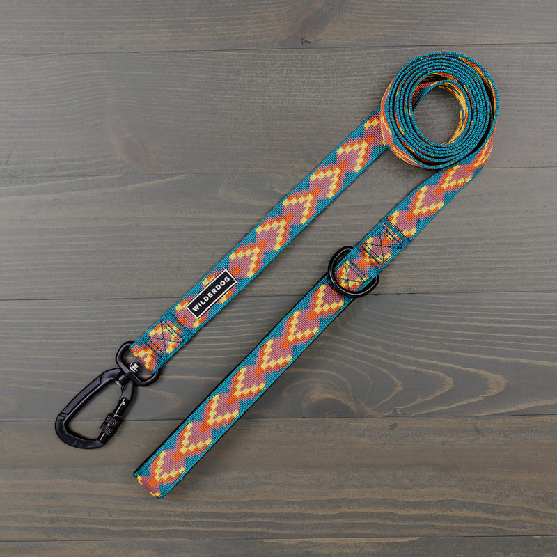 The Havasu Ultralight Leash, in a vibrant zigzag pattern of blue, orange, and yellow, stands out with its bright colors. Made of durable nylon webbing, it includes a black metal clip and a D-ring near the handle. The 'Wilderdog' brand name is displayed on a tag attached to this ultralight leash, all presented on a wooden surface.